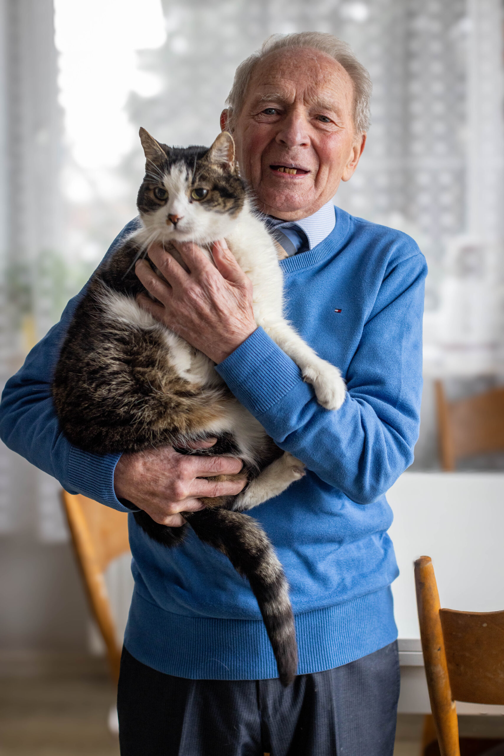 Starszy pan dziewięćdziesięciolatek w niebieskim swetrze uśmiechnięty trzyma na rękach kota