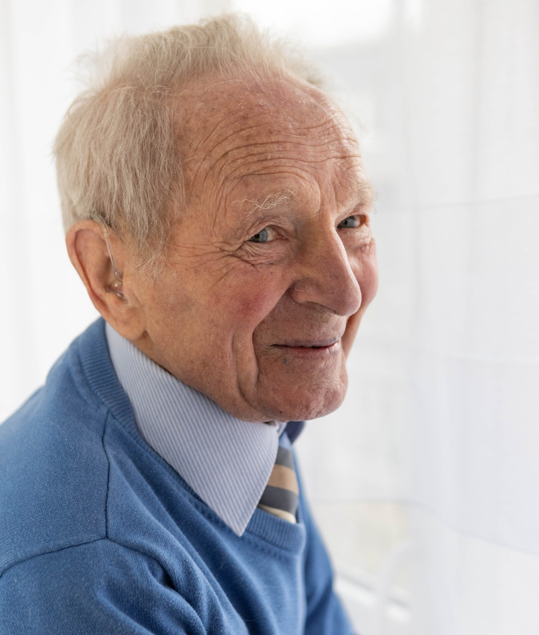 Starszy pan dziewięćdziesięciolatek uśmiechnięty pozuje przy oknie w niebieskim swetrze