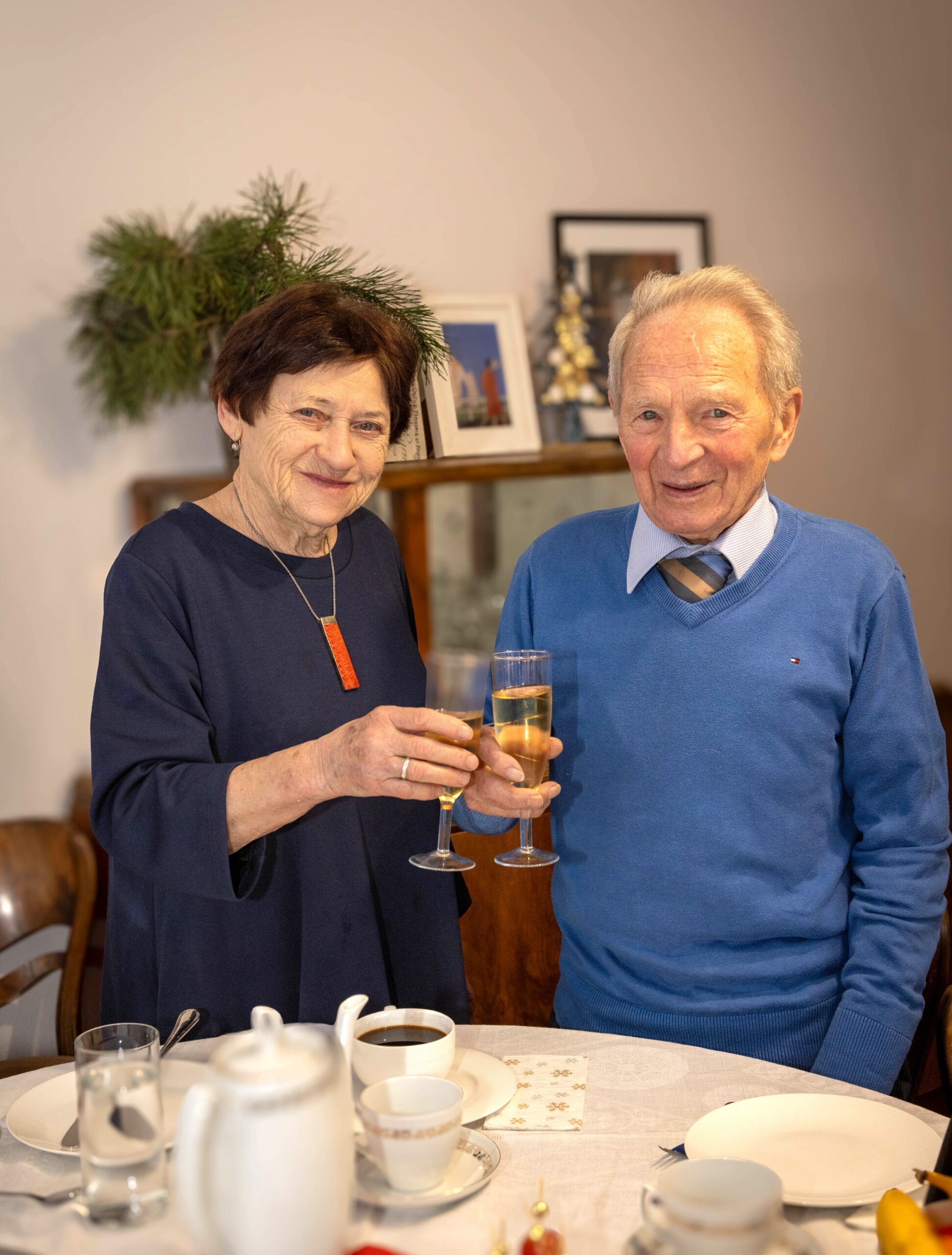 Starszy pan obchodzący dziewięćdziesiąte urodziny przy stole z żoną, trącają się kieliszkami z szampanem, sesja zdjęciowa dla seniorów prezent na dzień babci i dziadka, rodzinna sesja zdjęciowa babcia dziadek fotograf Jagoda Rogoża