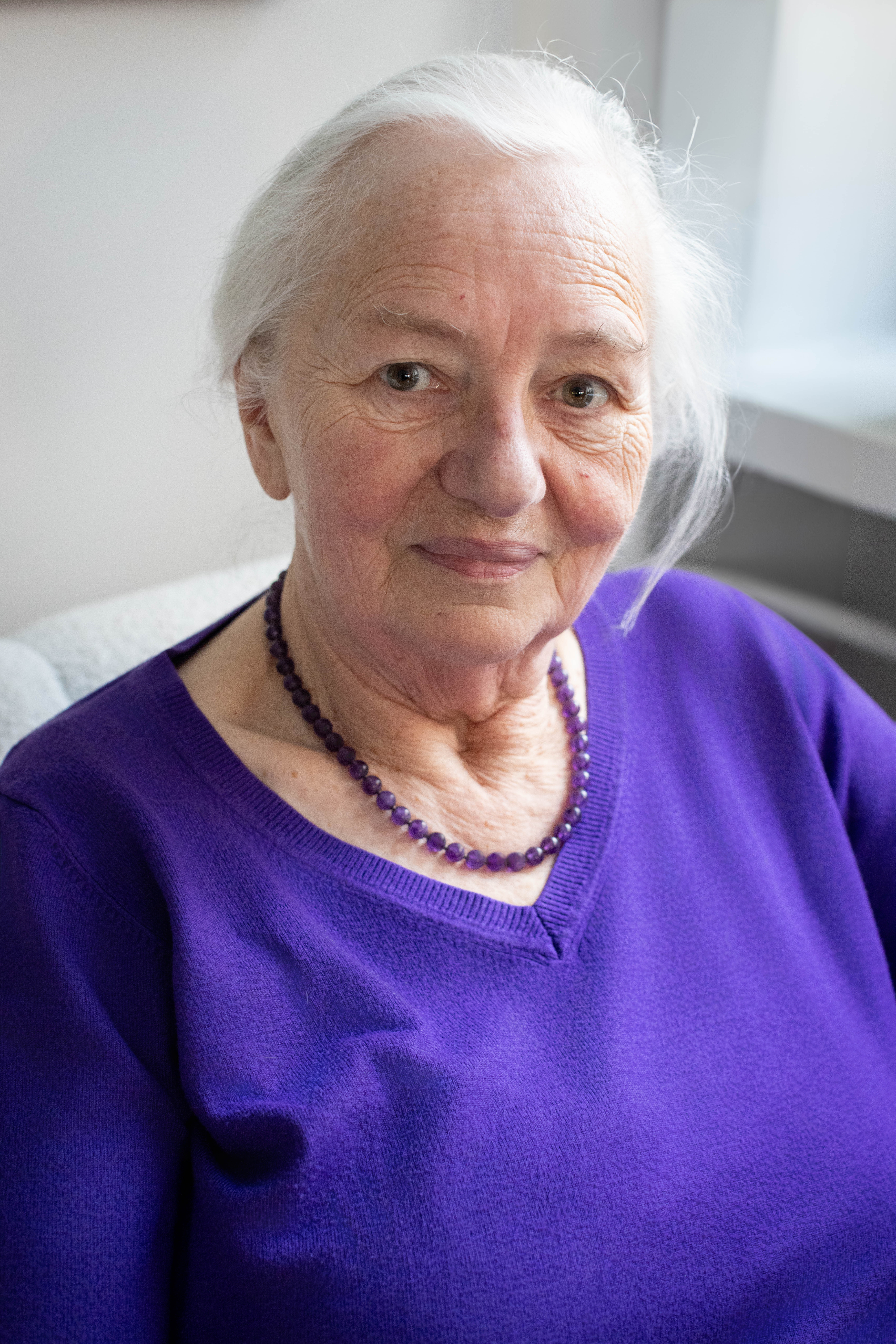 Starsza siwa pani we fioletowym sweterku z uśmiechem spogląda w obiektyw