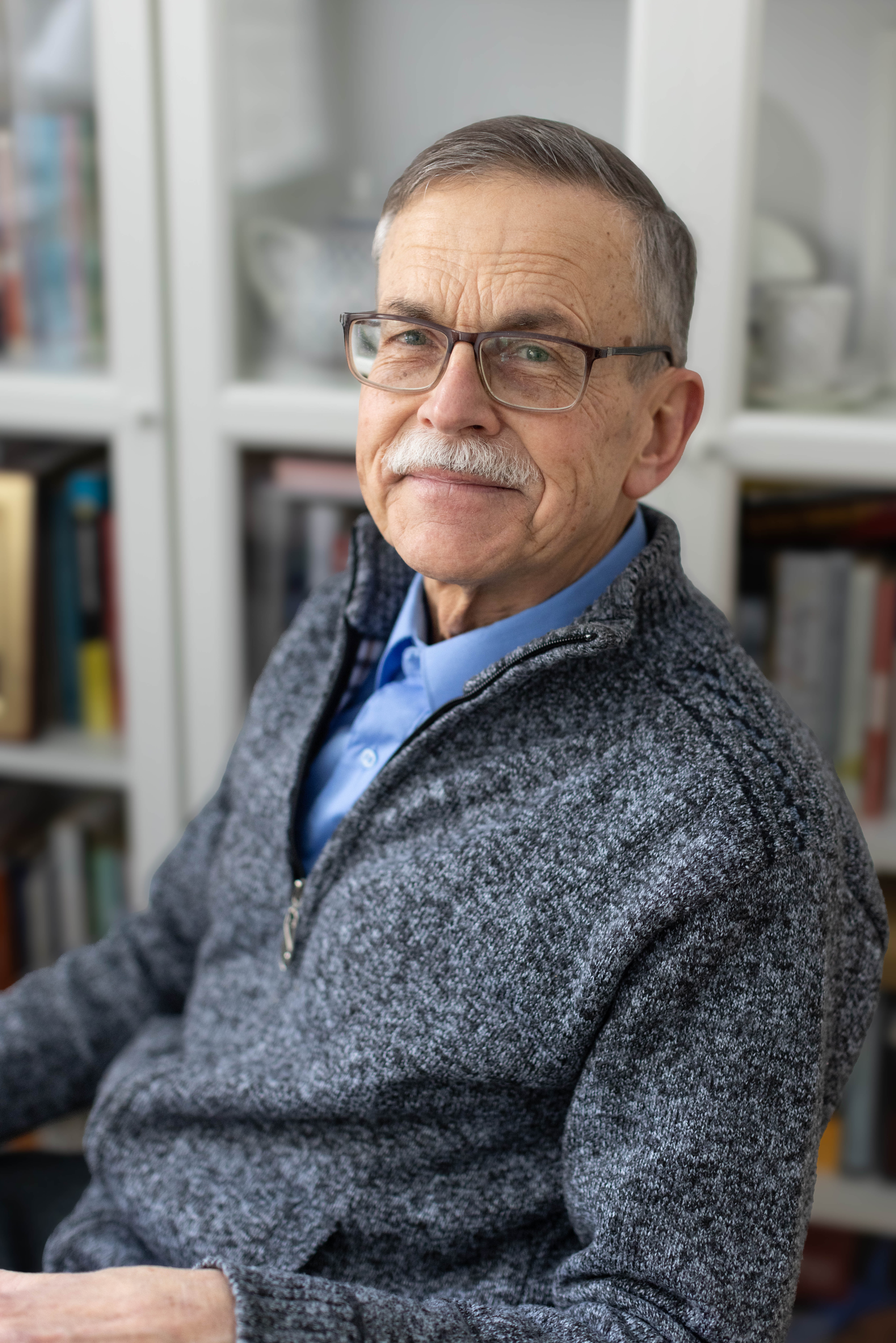 Starszy pan w okularach z siwym wąsem uśmiecha się, patrząc w obietkyw