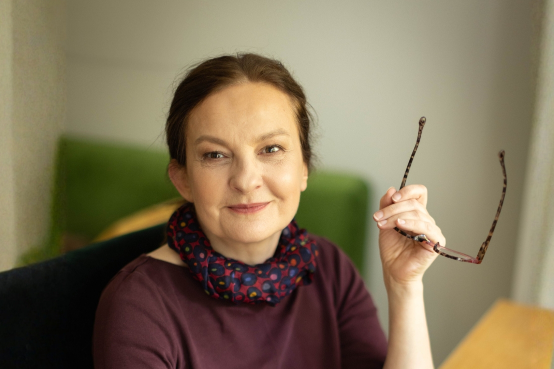 Portret rudowłosej kobiety trzymającej w ręku okulary i patrzącej z uśmiechem w obiektyw