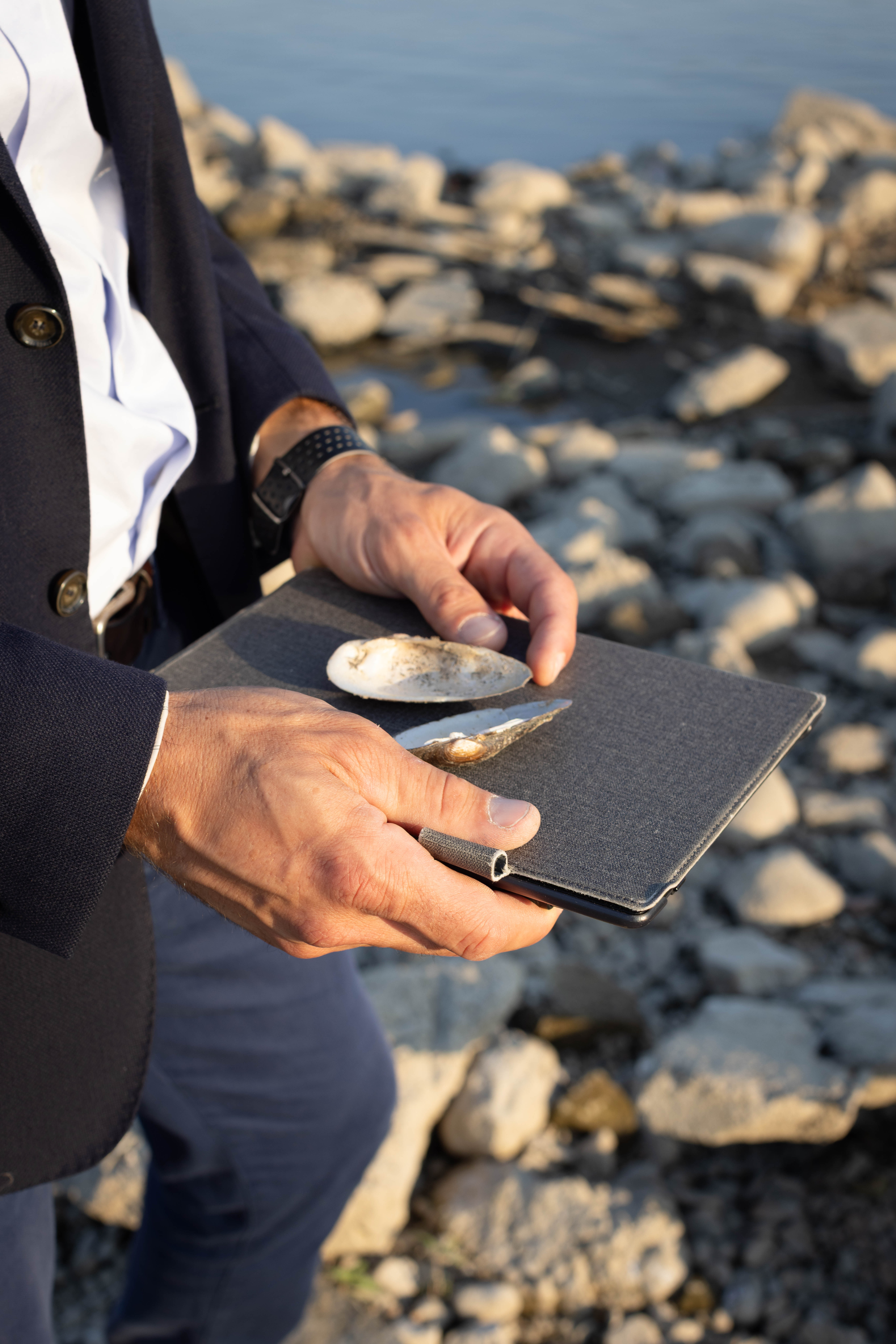 Detal, ręce mężczyzny w garniturze trzymają tablet i muszle, w tle kamieniste nabrzeże rzeki