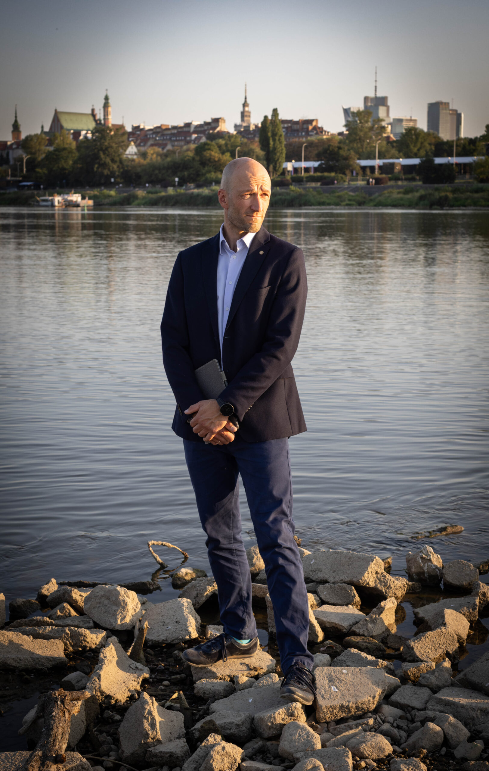 Mężczyzna w marynarce nad brzegiem rzeki, na tle warszawskiej starówki