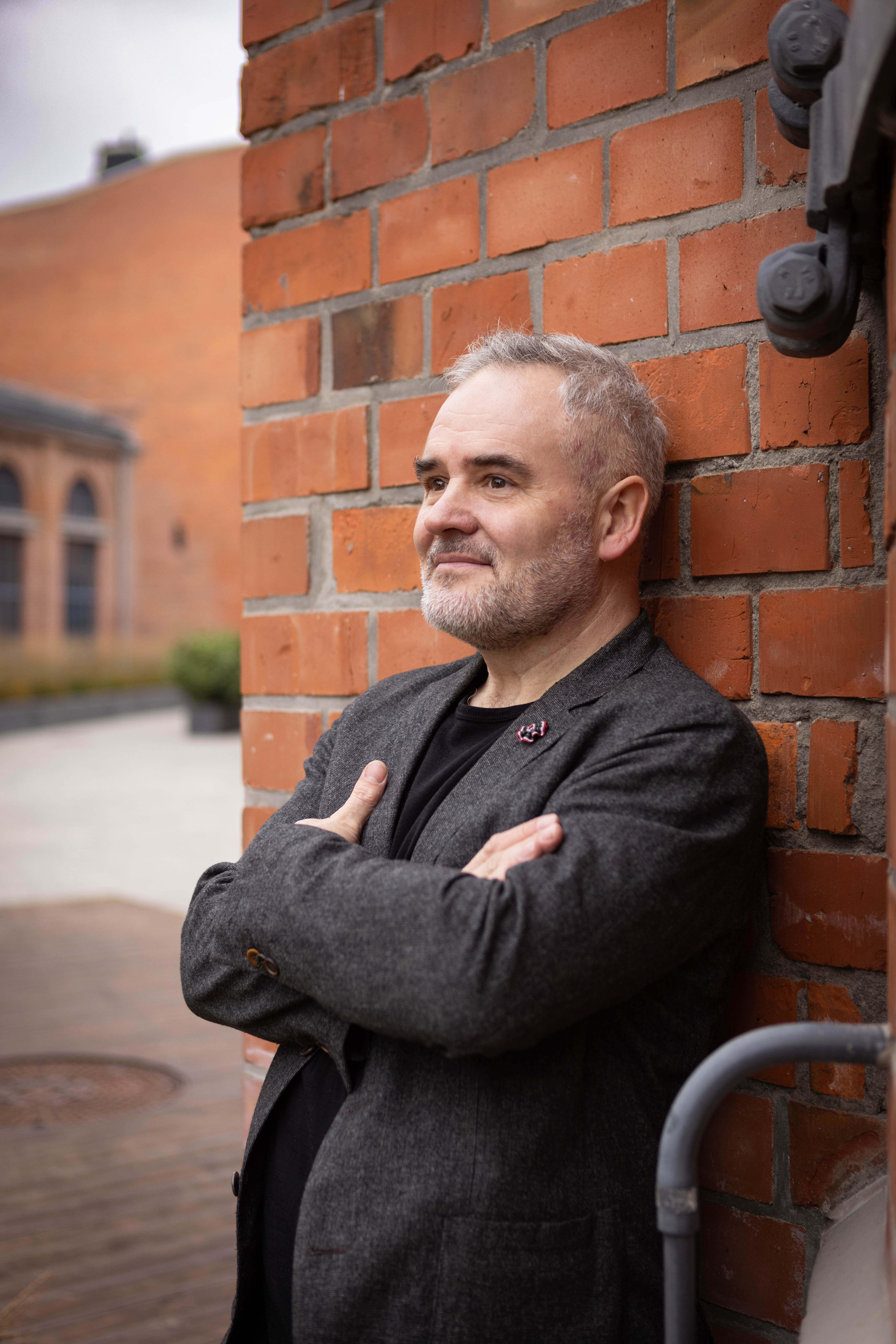 Mężczyzna w marynarce z założonymi rękami na tle ceglanej ściany