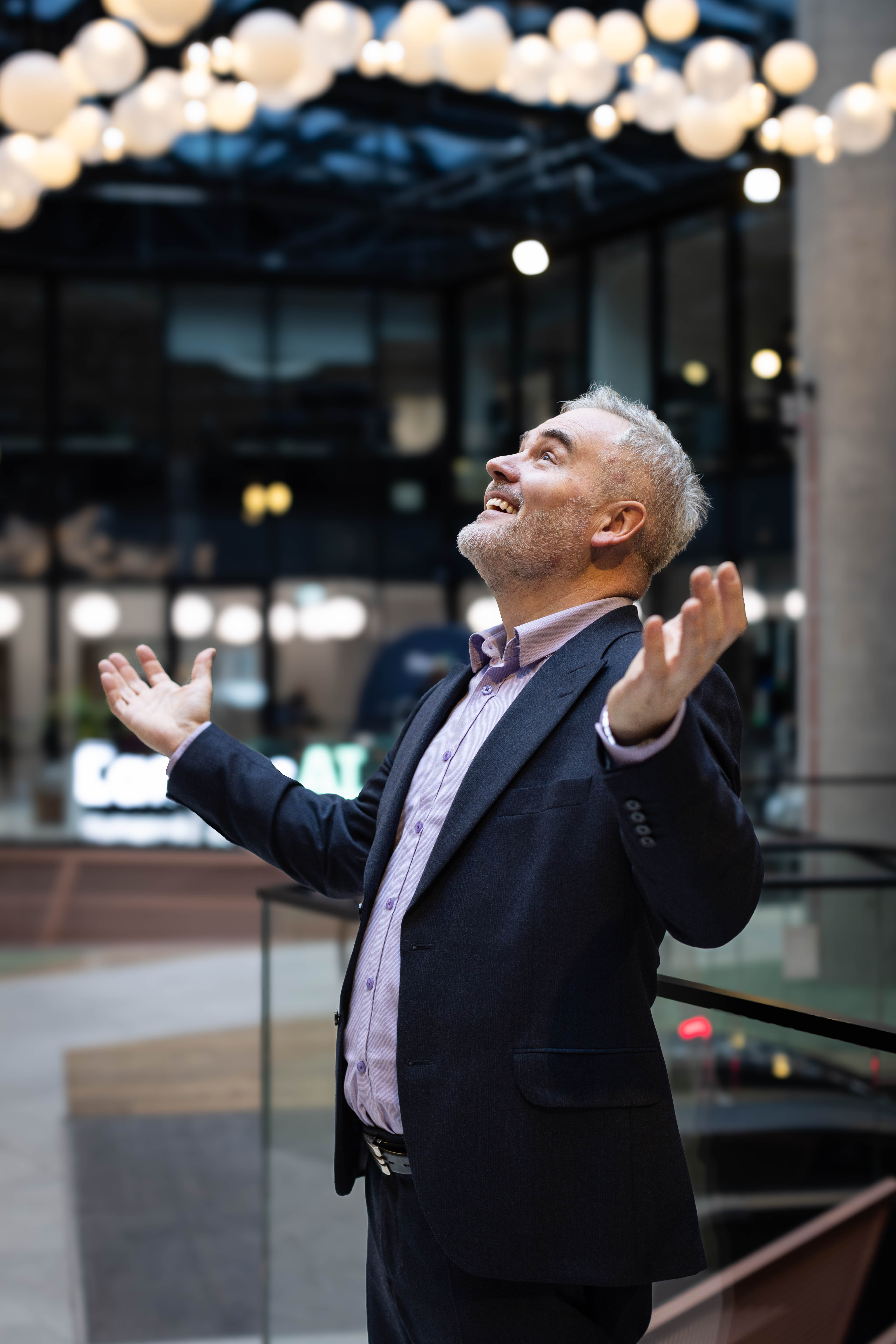 Mężczyzna w garniturze z podniesionymi rekami uśmiecha się, a nad nim są lampy