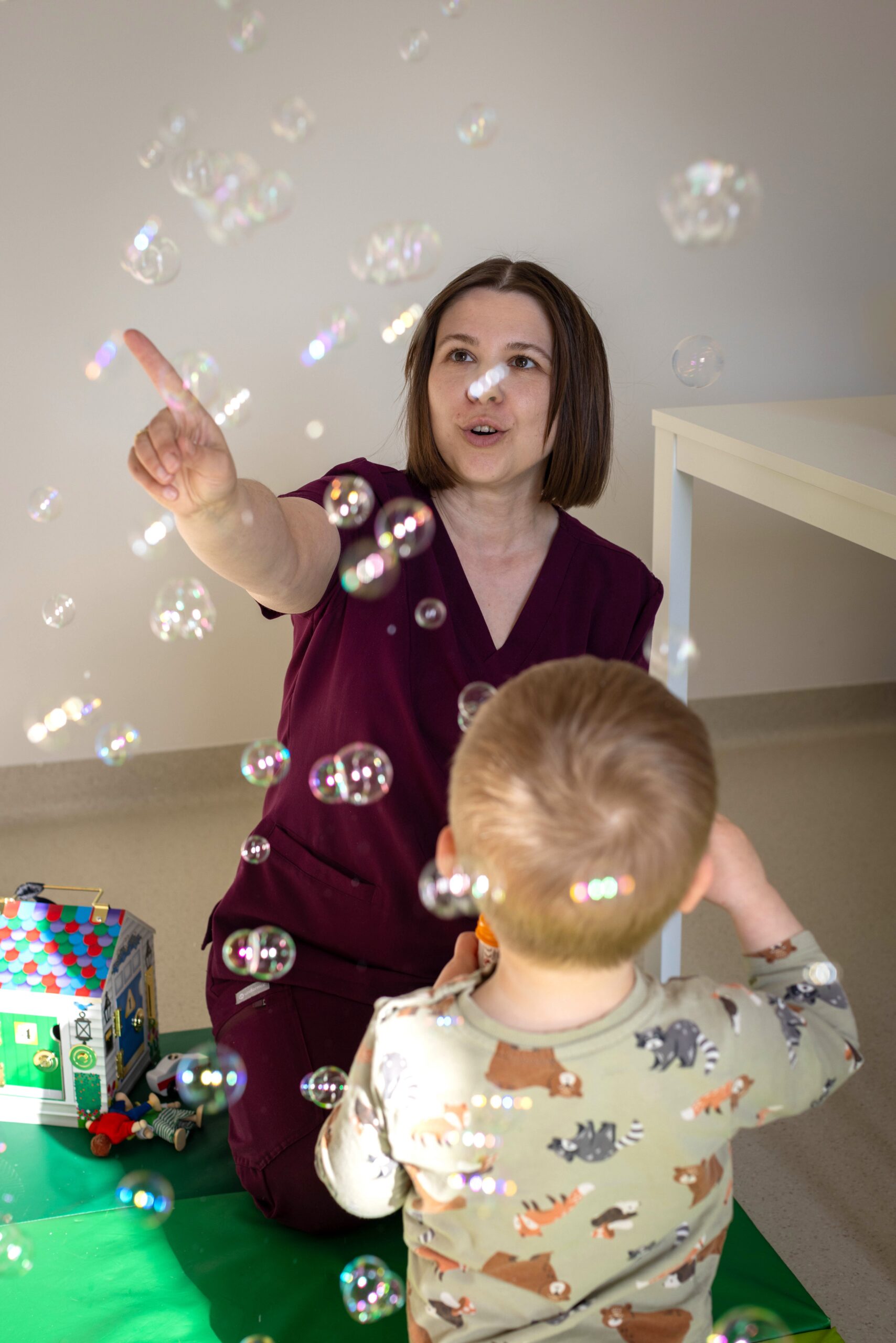 młoda śniada kobieta w bordowym kombinezonie roboczym bawi się z dzieckiem puszczając bańki mydlane